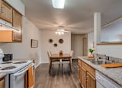 Thumbnail 22 of 47 - Spokane, WA, Apartments - Canyon Bluffs - Fully Equipped Kitchen with Appliances, Granite Countertops, Wooden Cabinetry, Wood-Style Flooring, Dual Sink, and View of Dining Area with a Table and Chairs.