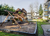 Thumbnail 29 of 35 - Two-Bedroom Apartments in Everett, Washington - Woodbrook - Outdoor Playground With A Wooden Play Structure, Swings, Slide, Mulch Ground Cover, Green Lawn, Trees, And Nearby Apartment Buildings.