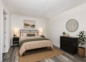 Thumbnail 6 of 23 - A bedroom with a large bed, a nightstand, a lamp, a plant, and a round mirror on the wall.