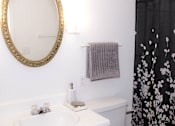 Thumbnail 17 of 17 - A bathroom with a white toilet, a white sink, a gold framed mirror, and a black and white shower curtain.