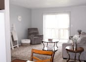 Thumbnail 5 of 17 - A living room with a grey couch, a brown chair, a grey armchair, a white ottoman, a small table, and a window with white curtains.
