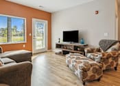 Thumbnail 4 of 24 - A living room with a grey sofa, a brown armchair, and a patterned ottoman.