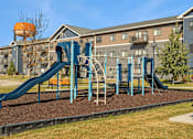 Thumbnail 18 of 24 - A playground with a blue slide and a brown sandbox in front of a building.