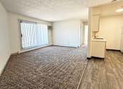Thumbnail 9 of 21 - A room with a carpeted floor, a window with blinds, a small kitchenette, and a door leading to another room.