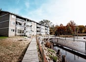 Thumbnail 16 of 18 - A wooden walkway leads to apartment buildings by a small body of water.