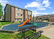 Thumbnail 14 of 26 - A playground area with a slide and picnic table in front of apartment buildings.