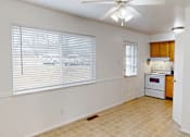 Thumbnail 6 of 16 - A room with a fan, a window with blinds, and a kitchen area with a stove and oven.