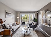 Thumbnail 3 of 31 - A living room with a grey couch, a glass coffee table, a grey armchair, a grey side table, a grey cabinet, a round mirror, a framed picture, a potted plant, a window with curtains,