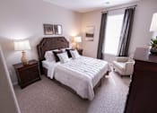 Thumbnail 14 of 20 - A bedroom with a large bed, a nightstand with a lamp, a chair, and a window with curtains.