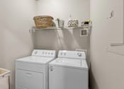 Thumbnail 11 of 19 - A laundry room with two washing machines and a shelf with various items on it at Heights at New Garden apartments in Greensboro, NC.