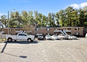 Thumbnail 48 of 52 - A parking lot with cars and a truck in front of a the exterior brick building with trees in the background.