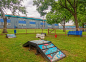 Thumbnail 19 of 25 - The Canvas apartments dog park with agility course, pet waste station, large green trees, and apartment buildings in the background.