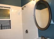 Thumbnail 13 of 29 - Bathroom in one bedroom brookmore apartment in Pasadena. Shower with white tile and silver fixtures and open window, and oval mirror above pedestal sink