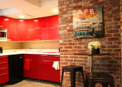 Thumbnail 20 of 29 - Brookmore apartments community kitchen with red cabinets, dishwasher, and microwave, and sitting area with small bar table and two black stools.