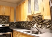 Thumbnail 11 of 29 - A kitchen in Brookmore apartments in Pasadena ca with modern, honey wood cabinets, multicolored tile backsplash, six burner gas stove, and dual sink