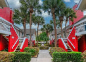 Thumbnail 49 of 60 - Fusion Orlando apartments exteriors with red staircases, blue front doors, and white railing on balconies. Center path is lined with palm trees.