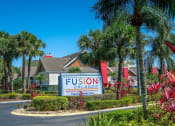 Thumbnail 46 of 60 - Main entrance to Fusion furnished apartments in Orlando, FL. Palm trees, bushes, and tropical flora line the main road and median with Fusion Orlando sign.
