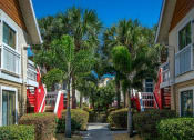 Thumbnail 44 of 60 - Fusion Orlando apartments pathway between buildings lined with lush bushes and palm trees. Red staircases lead up to second story.