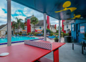Thumbnail 33 of 60 - Fusion Orlando apartments community pool next to red ping pong table under awning with bright yellow ceiling fans.