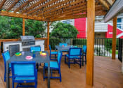 Thumbnail 32 of 60 - Fusion Orlando apartments outdoor grilling area with tables and chairs, grill and prep counters, and trash can under stained wood trellis.