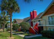 Thumbnail 47 of 60 - Street-facing Fusion Orlando apartments. Palm trees and a black iron fence line the main road and concrete paths lead to apartment doors.