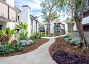 Thumbnail 10 of 15 - Fusion Warner Center luxury apartments in Woodland Hills, CA pathway between two-story buildings with lush green landscaping and brown bark mulch.