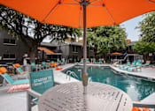 Thumbnail 17 of 36 - Large community pool and poolside table and chairs with orange umbrella at Stadium 700 apartments in Arlington, TX