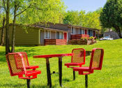 Thumbnail 39 of 49 - The Retreat at Indian Lake grounds with red steel mesh picnic table and chairs on green lawn with apartments in background.