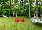 Thumbnail 32 of 36 - A red chair and a blue and white striped hammock are set up in a grassy area with trees in the background at the Retreat at Indian Lake Apartments.