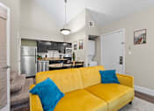 Thumbnail 3 of 41 - A yellow couch with a blue pillow is in the foreground of a kitchen and living room at Fusion Orlando furnished apartments.
