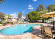 Thumbnail 14 of 15 - Fusion Warner Center luxury apartments in Woodland Hills pool with tan lounge chairs and patio tables with tan umbrellas on natural stone tile ground.