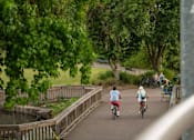 Thumbnail 5 of 8 - A man and a woman ride bicycles on a bridge.