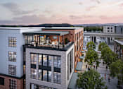 Thumbnail 1 of 8 - A modern building with a glass facade is surrounded by trees and other buildings.