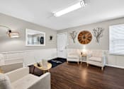 Thumbnail 6 of 31 - A living room with a grey couch, a white armchair, a wooden coffee table, and a large clock on the wall.
