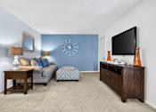 Thumbnail 7 of 31 - A living room with a grey couch, a wooden table, a blue ottoman, a clock, a flat screen TV, and a vase.