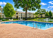 Thumbnail 5 of 15 - A swimming pool surrounded by a brick patio and a black fence with a building in the background.