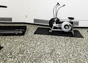 Thumbnail 16 of 16 - A stationary bike is on a black mat in a room with a white wall.