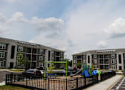 Thumbnail 13 of 22 - A playground with a slide and a green climbing frame is in front of apartment buildings.