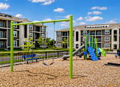 Thumbnail 11 of 22 - A playground with a green swing set and a blue slide in front of apartment buildings.
