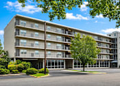 Thumbnail 1 of 6 - A large apartment building with a parking lot in front.