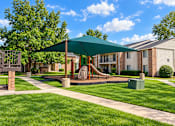 Thumbnail 8 of 33 - A playground with a green canopy and a slide is surrounded by a grassy area and apartment buildings.