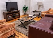 Thumbnail 11 of 25 - A living room with a brown leather couch, a patterned chair, a wooden cabinet, a glass coffee table, and a television.