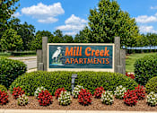Thumbnail 2 of 9 - A sign for Mill Creek Apartments is surrounded by flowers and greenery.