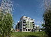 Thumbnail 1 of 7 - A modern apartment building with a grassy area in front.