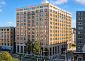 Thumbnail 2 of 8 - A large, beige building with many windows is in the foreground of a city street.