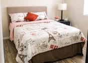 Thumbnail 2 of 9 - A bed with a white comforter and a pink pillowcase is in a room with a lamp and a picture frame on a nightstand.