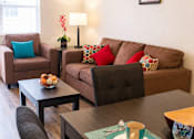 Thumbnail 5 of 9 - A living room with a brown couch, a brown chair, a brown armchair, a coffee table, a lamp, a vase with flowers, a bowl of fruit, a plate with a cup, and a picture on the wall