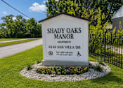 Thumbnail 1 of 12 - A sign for Shady Oaks Manor Apartments is displayed in front of a fence.