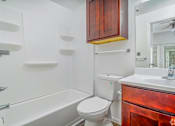 Thumbnail 14 of 25 - A white toilet sits next to a white bathtub in a bathroom with wooden cabinets.