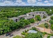 Thumbnail 6 of 25 - A large building with a grey roof is surrounded by greenery and a road with cars.